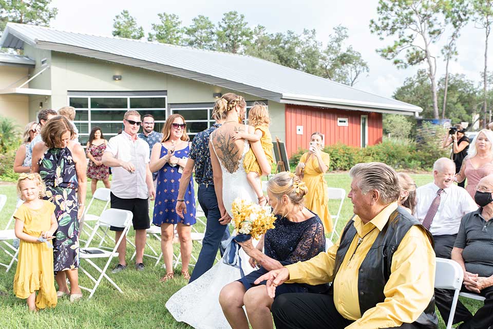 A destination wedding venue showcases an outdoor scene with a bride, adorned with a detailed back tattoo, holding a child as she walks down the aisle. Guests, in summer attire, are seated on white chairs with some standing. Lush green grass and a picturesque building complete the background.