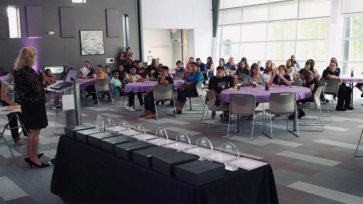 A speaker addresses an attentive audience seated at round tables during the Hernando Community Coalition Corporate Event, with awards displayed on a table at the front.