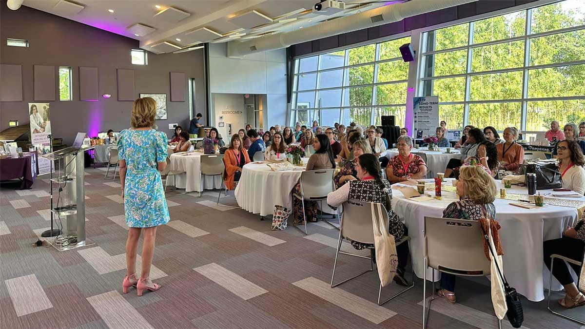 A conference speaker presents to an engaged audience at a corporate wellness event in a brightly lit, modern venue with nature visible through large windows.