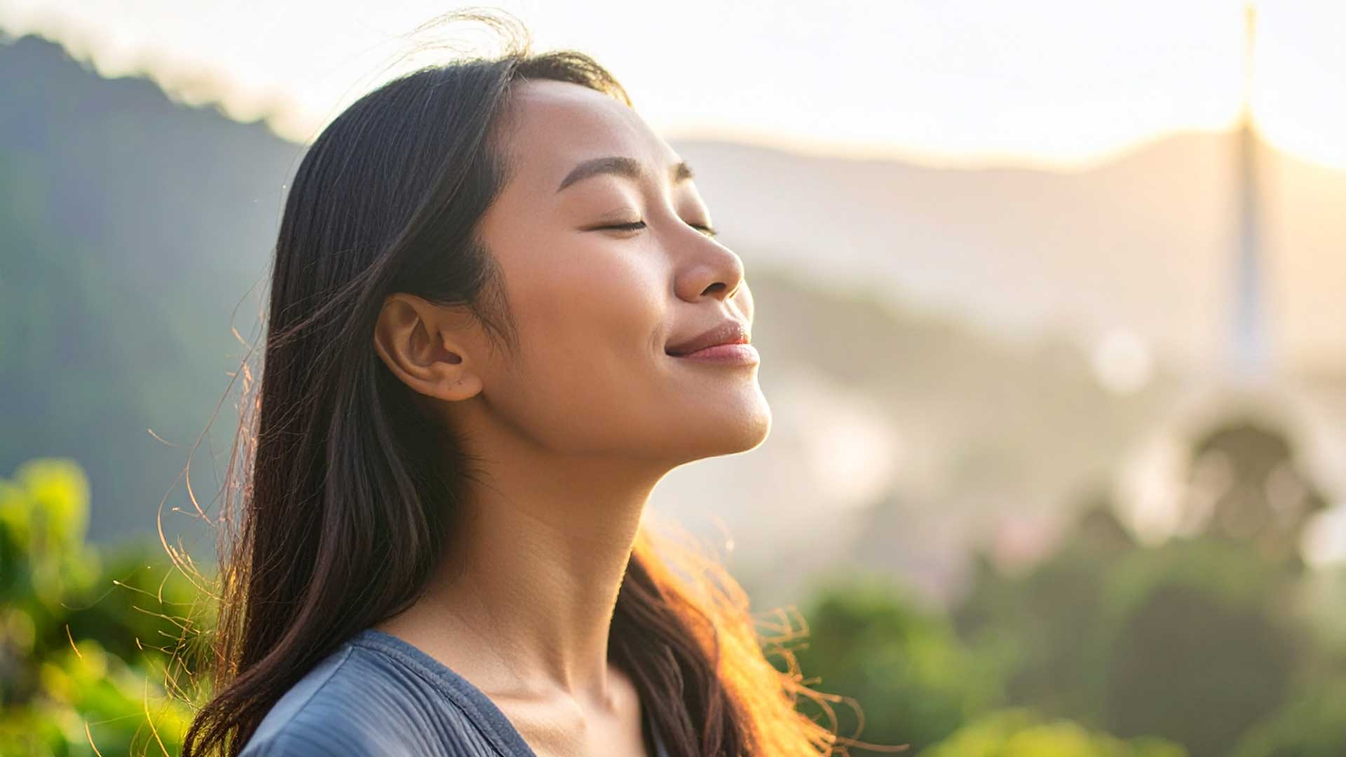 woman-air-toxins-nature-industry A serene woman standing outdoors in soft morning light, eyes closed, surrounded by invisible wisps or a subtle haze representing unseen toxins in the air. The background features both natural elements (trees, sky) and faint industrial cues (smokestack in the distance, power lines), symbolizing the contrast between nature and man-made environments. The mood is calm but thought-provoking.