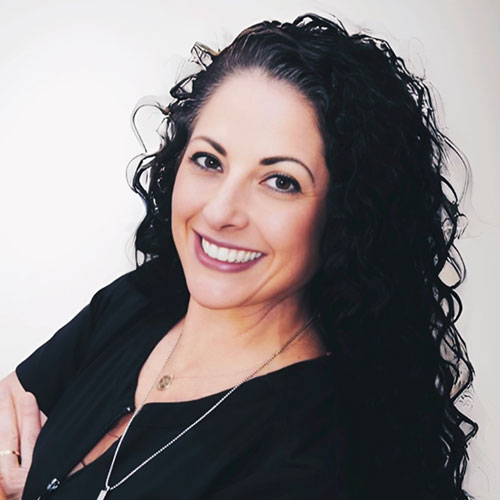 A woman with long, curly dark hair smiles at the camera. She is wearing a black top and a necklace, with a light-colored background behind her—radiating warmth often seen in massage therapy professionals.
