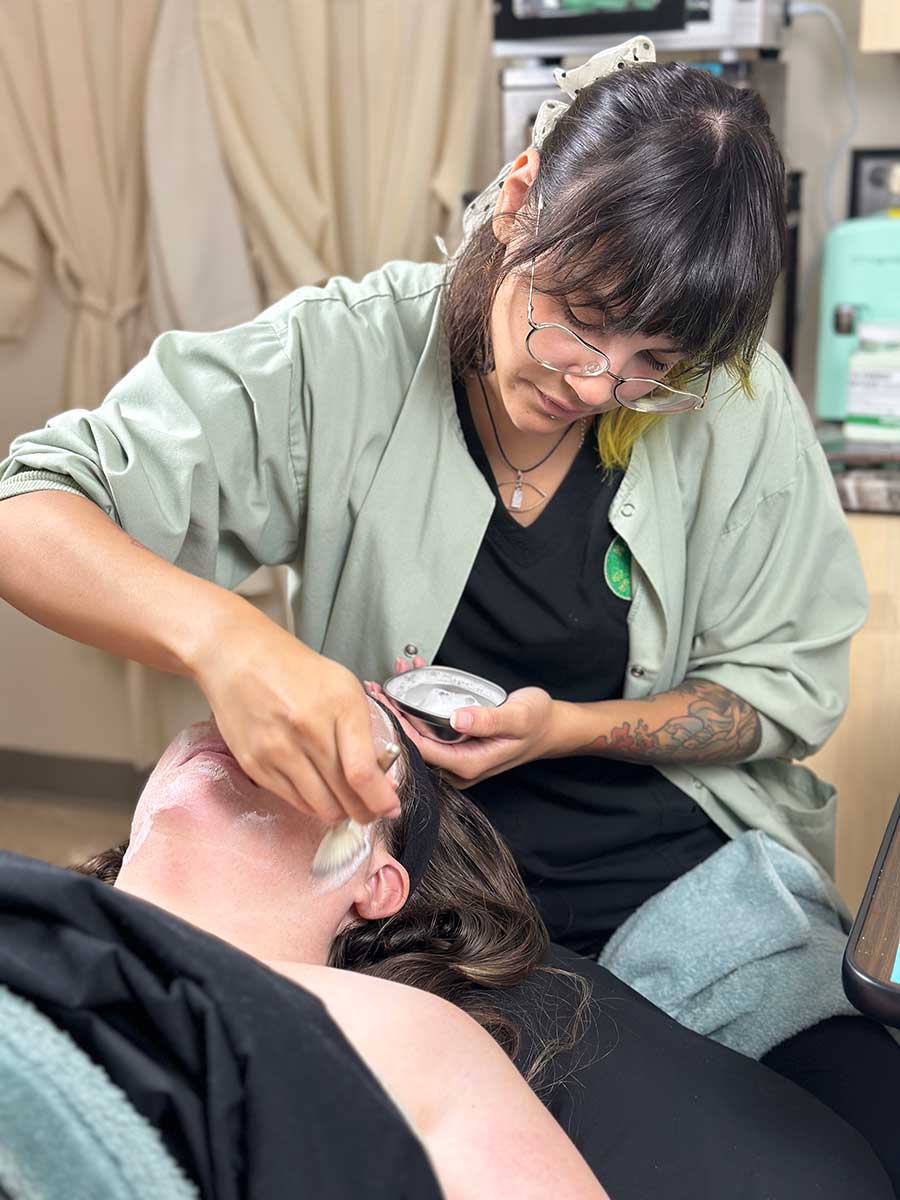 At Honey Beauty, a person wearing glasses and a green shirt carefully applies a facial treatment with a brush to another person lying down with a towel over their chest in the relaxing spa setting.