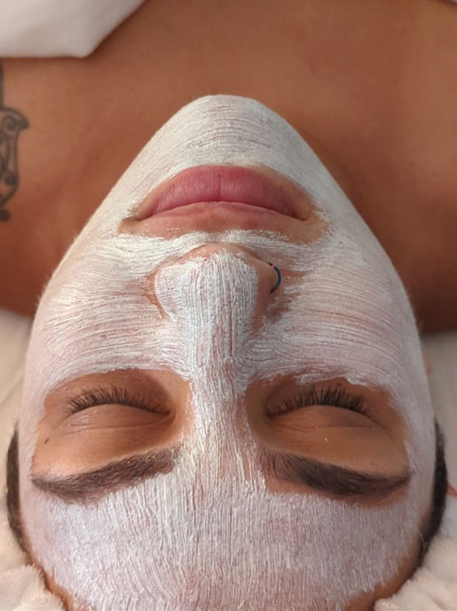 A person with closed eyes is lying down, covered with a white facial mask. A tattoo is visible on their shoulder, and they have a nose ring. The photo is taken from above, focusing on their face and relaxing expression.